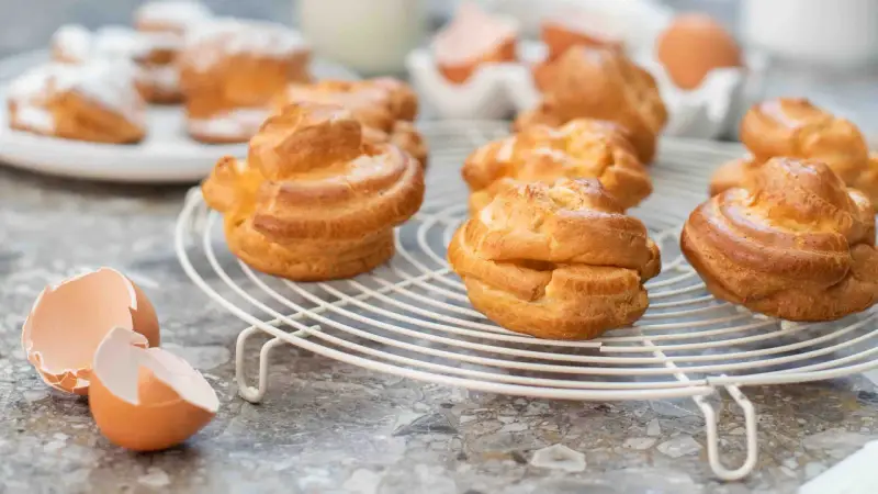Gyllene petit choux på ett vitt galler, med äggskal och ägg i bakgrunden.